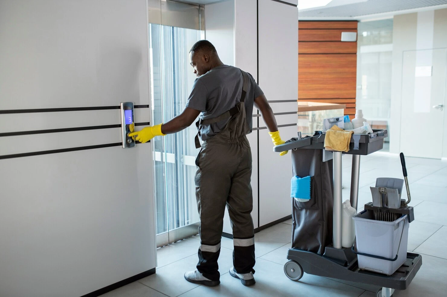 Cleaner pressing an elevator call button beside a fully stocked janitorial cart