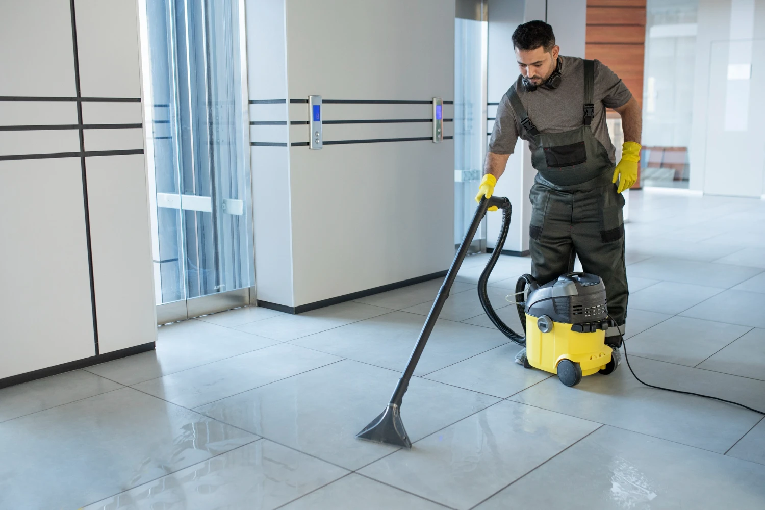 Cleaner vacuuming a wet tiled corridor near an elevator lobby